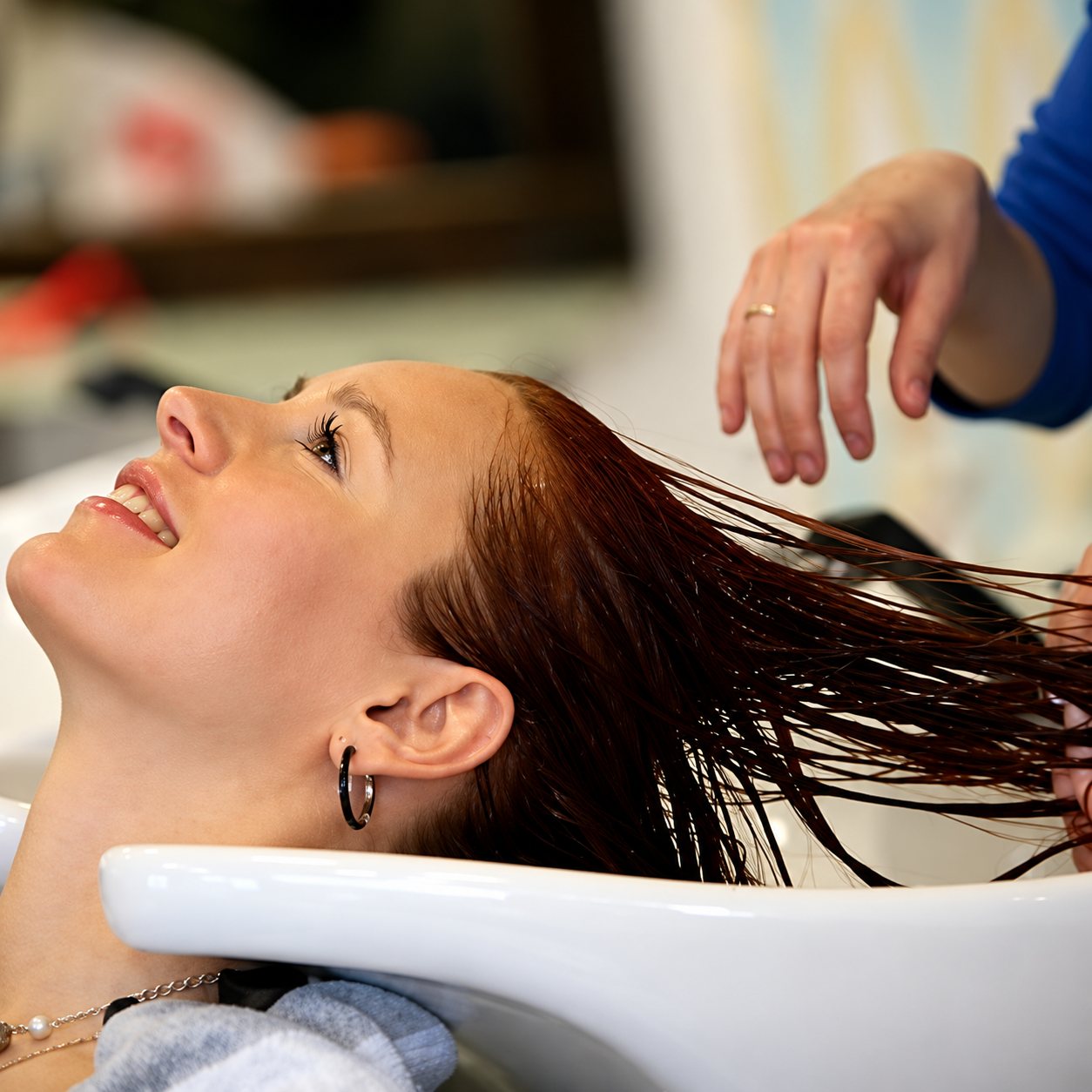 Une jeune femme aux cheveux auburn sourit, la tête renversée dans un bac de shampoing, tandis qu’une coiffeuse lui touche les cheveux.