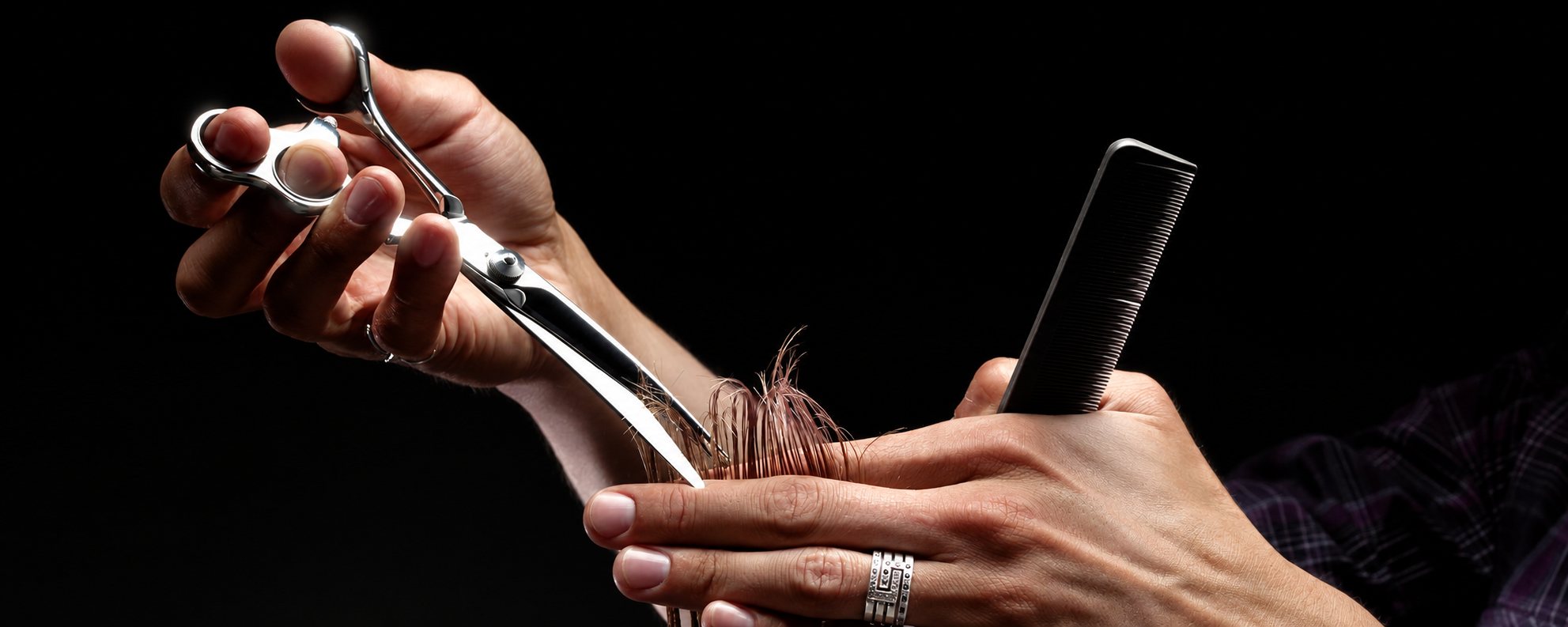 Deux mains de coiffeur tiennent des ciseaux et un peigne noir pour couper une mèche de cheveux châtains sur fond noir.